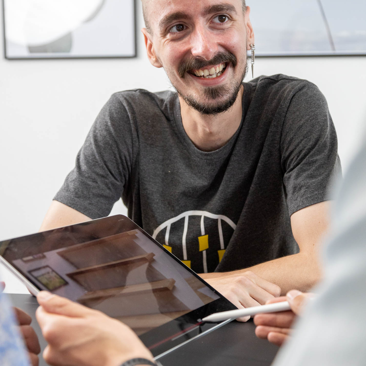 Ein lächelnder Mann mit Bart hält ein digitales Tablet und einen Stift in der Hand und interagiert mit jemandem außerhalb des Blickfelds während eines scheinbar gemeinschaftlichen Treffens bei einer Imagefilmagentur.