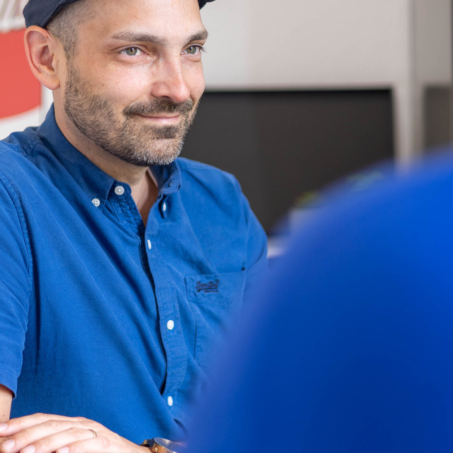 Der bärtige Mann in blauem Hemd und Mütze einer Imagefilmagentur hört einem Gespräch aufmerksam zu.